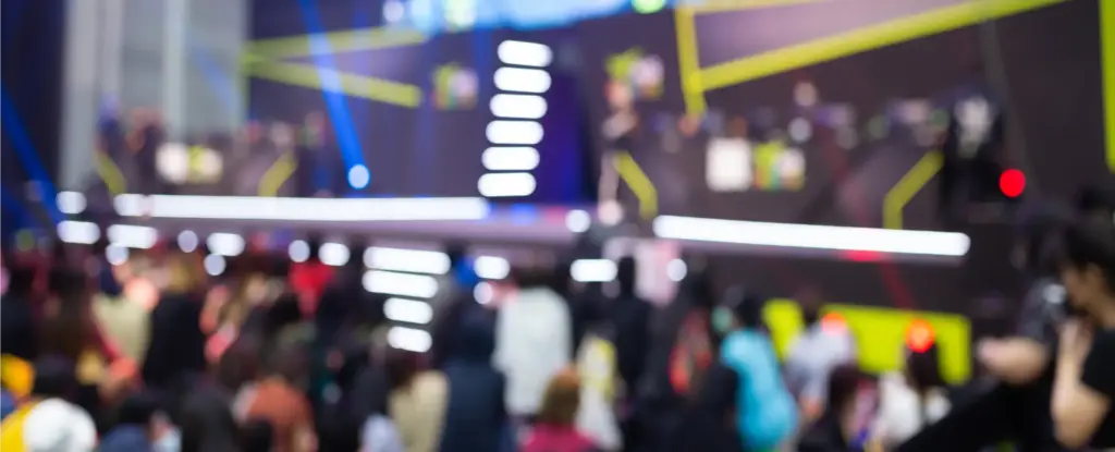 Een bruisend esports-evenement met een vervaagd podium en een groot scherm waarop beelden uit de game worden getoond. Een menigte kijkt vol verwachting toe, verlicht door kleurrijke lampen.