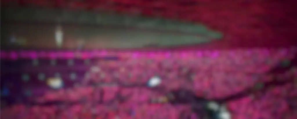 Een wazige foto van een grote concertzaal, gevuld met een zee van roze en paarse lichten. De sfeer is levendig en energiek, en er is een kronkelend podium te zien.