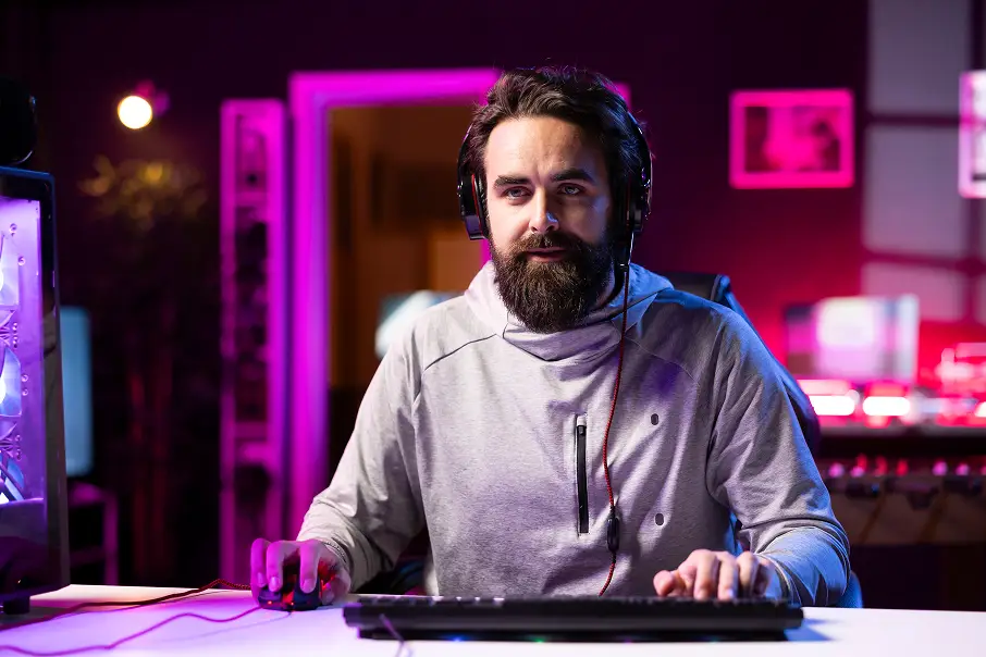 Een man met een koptelefoon zit te gamen op een computer, in een kamer die verlicht wordt door paarse en roze lampen. Hij ziet er geconcentreerd uit, wat een meeslepende, hightech sfeer creëert.