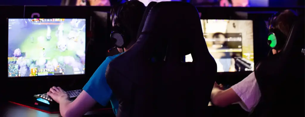 Drie gamers met een headset op staren geconcentreerd naar hun beeldschermen terwijl ze een videogame spelen. Een man in een wit overhemd houdt toezicht, waardoor er een competitieve, levendige sfeer ontstaat.