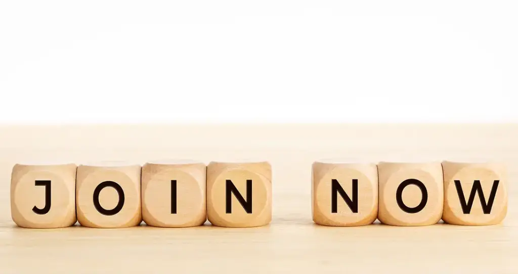 Op de houten blokjes staat in opvallende letters “JOIN NOW” afgebeeld op een lichtgekleurd houten oppervlak. De afbeelding roept op tot actie met een duidelijke, rechttoe rechtaan boodschap.