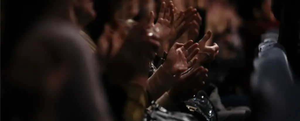 Vage beelden van een applaudisserend publiek in een slecht verlichte theaterzaal, die enthousiasme en waardering uitstralen. De handen en gezichten zijn enigszins onscherp, wat extra energie geeft.