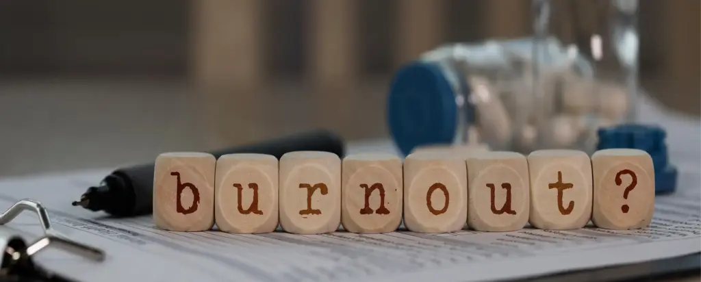 Houten blokjes vormen het woord “burn-out?” op een bureau met verspreide pillen, een receptbriefje en een pen, waarmee een thema van stress en uitputting wordt uitgedrukt.