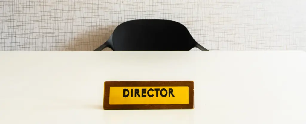 Een leeg bureau met een naamplaatje met het opschrift „Directeur“ erop, een zwarte stoel erachter, tegen een beige muur met structuur, wat een gevoel van professionaliteit en gezag uitstraalt.